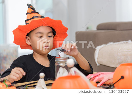Boy in a costume hat is focused on arranging Halloween decorations like spiders and ghosts around a jar, getting ready for a spooky celebration at home. Boy in a costume hat is focused on arranging Halloween decorations like spiders and ghosts around a jar, getting ready for a spooky celebration at home. 130433937