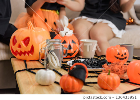 Children in costumes celebrate halloween day with jack o lanterns, spiders and a mason jar on a table decorated with a spiderweb, seated on a couch indoors for the spooky holiday. Children in costumes celebrate halloween day with jack o lanterns, spiders and a mason jar on a table decorated with a spiderweb, seated on a couch indoors for the spooky holiday. 130433951
