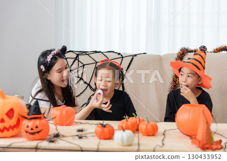 Festive family prepares for a halloween celebration, children in costume eating candy eyeballs and wearing hats sit near decorative pumpkins and spider webs. Festive family prepares for a halloween celebration, children in costume eating candy eyeballs and wearing hats sit near decorative pumpkins and spider webs. 130433952