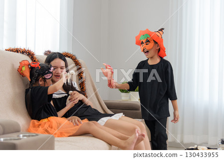 Woman lying with children on a sofa, decorations are on the sofa for halloween day, a child is wearing a mask, holding a fake hand, and playing with the siblings on holiday. Woman lying with children on a sofa, decorations are on the sofa for halloween day, a child is wearing a mask, holding a fake hand, and playing with the siblings on holiday. 130433955