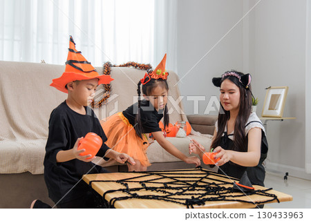Children in costumes enjoy halloween decorations, placing pumpkins and spiderwebs on a table, preparing for trick or treat and the festive season in a cozy home setting. 130433963