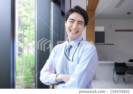 一位精神抖擻的年輕人，穿著領帶和襯衫，雙臂交叉，站在窗邊的新鮮綠植旁，看著鏡頭，在商務環境中微笑 130434802