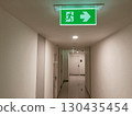 Illuminated green emergency exit sign with running man symbol and arrow pointing right, mounted on ceiling in modern building corridor, safety guidance for evacuation and emergency preparedness 130435454