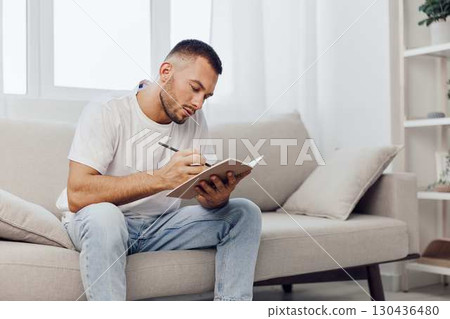 Thoughtful man writing in a notebook at home, wearing casual clothes, surrounded by natural light, reflecting on ideas and creativity. Concept: mindfulness. Thoughtful man writing in a notebook at home, wearing casual clothes, surrounded by natural light, reflecting on ideas and creativity. Concept: mindfulness. 130436480