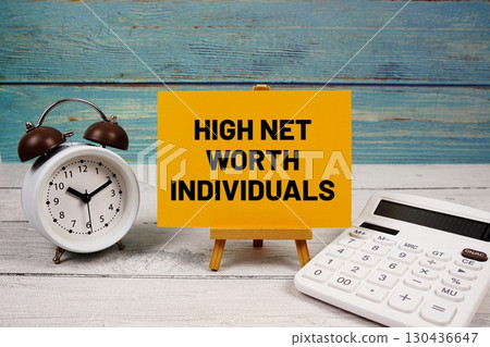 High Net Worth Individuals text written on paper card with calculator and alarm clock on wooden background 130436647