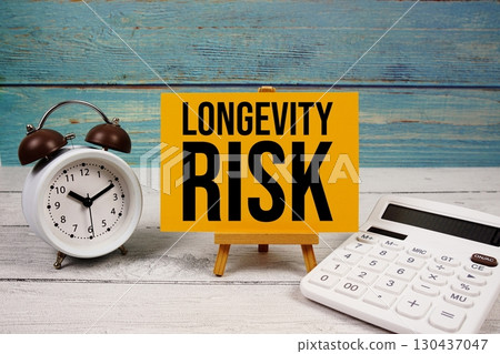 Longevity Risk text written on paper card with calculator and alarm clock on wooden background 130437047