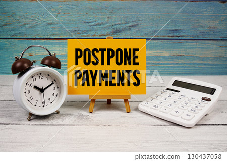 Postpone Payments text written on paper card with calculator and alarm clock on wooden background 130437058