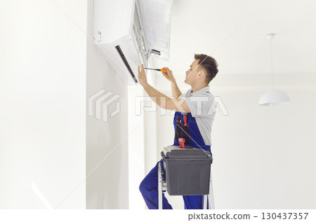 Professional electrician man with screwdriver maintaining modern air conditioner indoors. Professional electrician man with screwdriver maintaining modern air conditioner indoors. 130437357