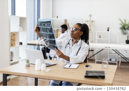 Confident young adult female doctor evaluating X-ray scans in office workspace. Medical professional focusing on diagnostics and patient records. Modern healthcare settings emphasizing professionalism Confident young adult female doctor evaluating X-ray scans in office workspace. Medical professional focusing on diagnostics and patient records. Modern healthcare settings emphasizing professionalism 130438762