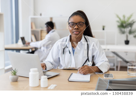 Confident young woman doctor using laptop and writing prescription in modern medical office. Bright workspace featuring healthcare equipment, teamwork depicted with colleague in background 130438839
