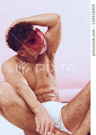Stylish young man with curly hair wearing pink visor and white briefs, posing against soft pink background. Modern fashion and self-expression concept. Stylish young man with curly hair wearing pink visor and white briefs, posing against soft pink background. Modern fashion and self-expression concept. 130438909