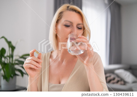Blonde woman looking at pill in hand thoughtfully, glass of water in the other hand sitting in modern interior Blonde woman looking at pill in hand thoughtfully, glass of water in the other hand sitting in modern interior 130439611