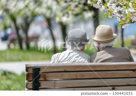 An elderly couple sits close together on a park bench under blooming trees, enjoying a peaceful spring day. They wear hats and seem to be engaged in a quiet, intimate conversation 130441031