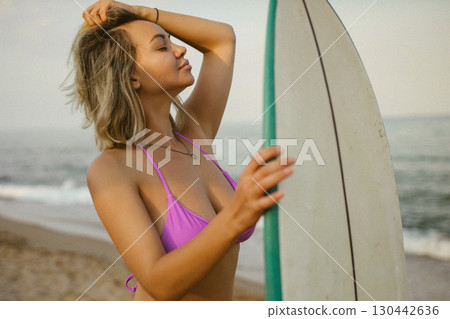 Attractive young woman in a purple bikini posing with a surfboard on the beach, enjoying summer by the sea, confident and relaxed, ocean waves in the background. 130442636