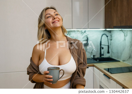 Modern kitchen: Young woman in a white crop top and open shirt stands with a mug in hand, enjoying a peaceful morning at home, relaxed and smiling. Modern kitchen: Young woman in a white crop top and open shirt stands with a mug in hand, enjoying a peaceful morning at home, relaxed and smiling. 130442637