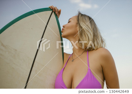 Blonde woman in purple bikini playfully kisses surfboard on sandy beach, summer sunset atmosphere, joyful and carefree vacation mood. Blonde woman in purple bikini playfully kisses surfboard on sandy beach, summer sunset atmosphere, joyful and carefree vacation mood. 130442649
