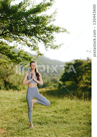Young woman practicing yoga in nature, wearing gray activewear, embodying tranquility and mindfulness amidst green landscapes 130443600