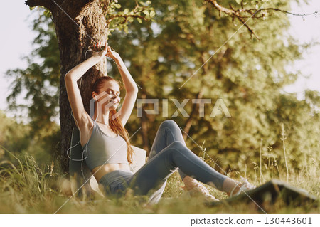 A young woman with long hair enjoying music while sitting under a tree in a sunny outdoor setting, wearing casual clothes, conveying relaxation and happiness A young woman with long hair enjoying music while sitting under a tree in a sunny outdoor setting, wearing casual clothes, conveying relaxation and happiness 130443601