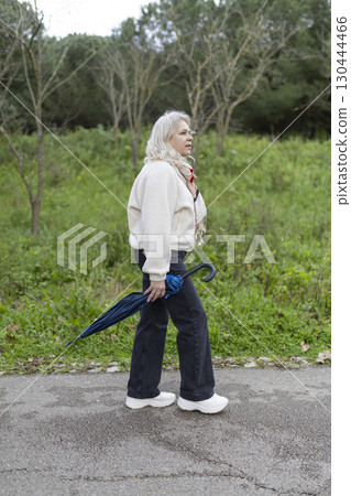 Senior woman walking in park holding closed umbrella on cloudy day 130444466