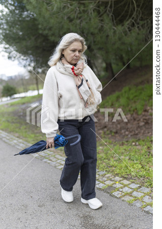 Sad senior woman walking in park after rain holding closed umbrella Sad senior woman walking in park after rain holding closed umbrella 130444468