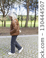 Senior woman walking in park wearing brown jacket and jeans 130444507