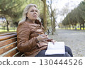 Senior woman thinking on park bench holding eyeglasses and notebook 130444526