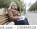 Senior woman writing notes in notebook while sitting on bench in park 130444528