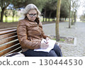 Senior woman writing in notebook while sitting on park bench 130444530