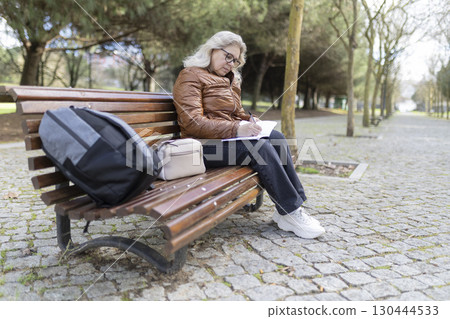 Senior woman writing notes in notebook while sitting on bench in park Senior woman writing notes in notebook while sitting on bench in park 130444533