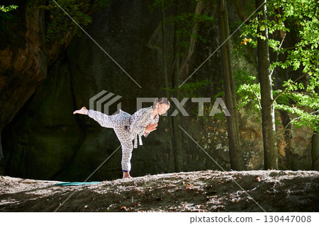 Woman practicing yoga outdoors in forest. Barefoot female on yoga mat surrounded by trees and large rocks, which suggests peaceful, natural environment ideal for meditation or yoga practice. Woman practicing yoga outdoors in forest. Barefoot female on yoga mat surrounded by trees and large rocks, which suggests peaceful, natural environment ideal for meditation or yoga practice. 130447008