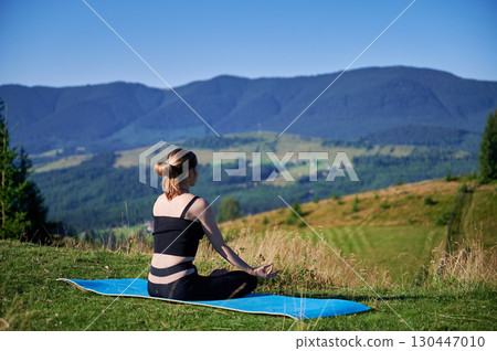 Woman practicing yoga and meditating outdoors in the mountains in serene, natural setting. Female performing yoga pose on blue mat, with backdrop of beautiful mountain landscape at sunrise or sunset. 130447010