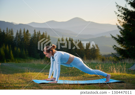 Woman practicing yoga outdoors in the mountains in a serene, natural setting. Female performing yoga pose on blue mat, with backdrop of beautiful mountain landscape at sunrise or sunset. 130447011