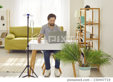 Young man working on a laptop at the desk at home or in office while receiving IV vitamin therapy. Young man working on a laptop at the desk at home or in office while receiving IV vitamin therapy. 130447719