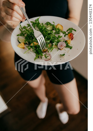 Woman wearing sportswear holding plate with fork and eating fresh healthy salad with arugula, cucumber, peaches and feta cheese, healthy lifestyle and eating habits concept. Wellness focused lifestyle 130448174
