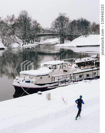 Man is jogging along snow covered riverbank, passing moored boat blanketed in snow, with bridge and snow laden trees in background during serene winter day. Male runs traversing snow covered riverbank Man is jogging along snow covered riverbank, passing moored boat blanketed in snow, with bridge and snow laden trees in background during serene winter day. Male runs traversing snow covered riverbank 130448183