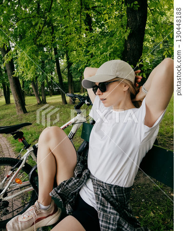 Young blonde woman cyclist wearing white t shirt, shorts, sneakers and cap is sitting on bench in green park after cycling, holding her hair with her hands and resting near her bicycle. Woman pausing Young blonde woman cyclist wearing white t shirt, shorts, sneakers and cap is sitting on bench in green park after cycling, holding her hair with her hands and resting near her bicycle. Woman pausing 130448188