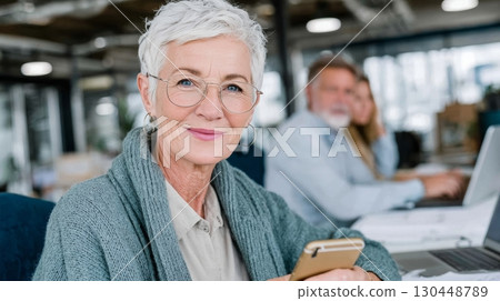 Senior woman with short gray hair is smiling while using smartphone in modern office, showcasing an ageless workforce and longevity at work 130448789