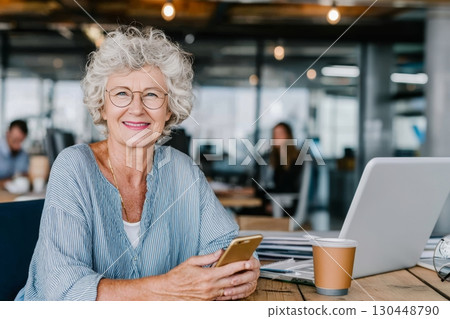 Senior woman with curly hair smiles while using smartphone at modern workspace, showcasing the ageless workforce and longevity at work 130448790