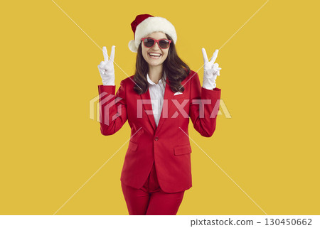 Happy woman wearing red suit, Santa Claus Christmas hat showing victory sign on yellow background. Happy woman wearing red suit, Santa Claus Christmas hat showing victory sign on yellow background. 130450662