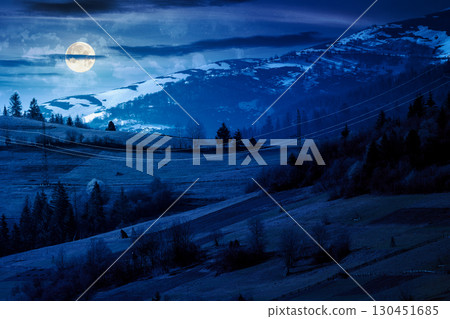 mountainous countryside landscape in spring at night. grassy field on slope of a hill. mountain ridge with snow capped tops in the distance in full moon light. sunny weather with clouds on the sky 130451685