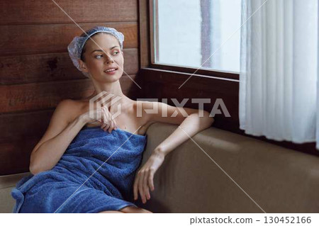Relaxed woman wrapped in a blue towel with a hair towel turban, sitting by a window in a wooden spa room, enjoying a peaceful moment of self-care and calmness. 130452166