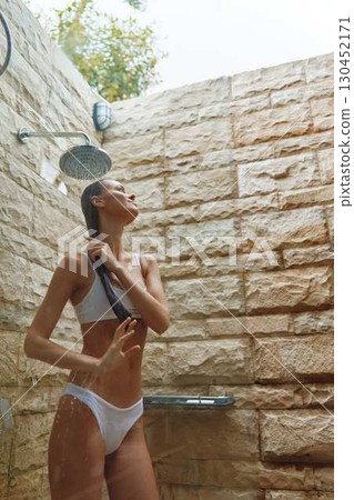 woman taking outdoor shower in white swimwear with wet hair, stone wall background, natural light, fresh and relaxed mood, summer wellness moment 130452171