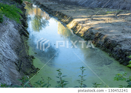 A tranquil waterway sits amidst a construction site, surrounded by empty land and lush greenery under clear blue skies 130452241