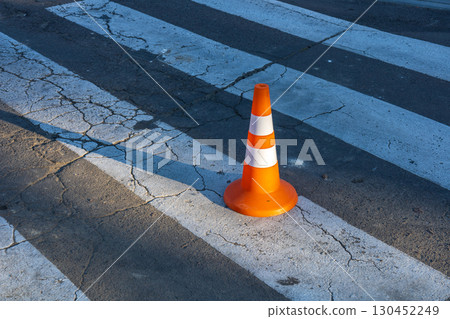 A traffic cone stands on a cracked crosswalk, casting shadows on the pavement in the morning light, indicating road work or caution A traffic cone stands on a cracked crosswalk, casting shadows on the pavement in the morning light, indicating road work or caution 130452249