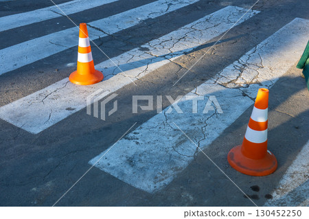 Bright orange traffic cones are positioned on a worn crosswalk, indicating construction or caution at a bustling city intersection in the evening 130452250