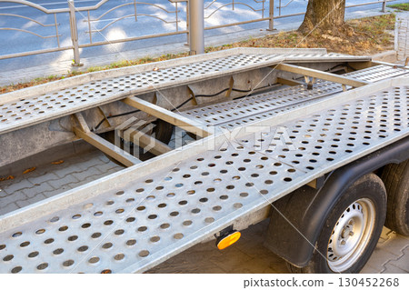 A ramp of a vehicle trailer rests on a paved surface in an urban setting, illuminated by sunlight and surrounded by greenery A ramp of a vehicle trailer rests on a paved surface in an urban setting, illuminated by sunlight and surrounded by greenery 130452268