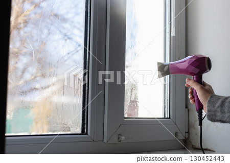 A womans hand drying wet window glass with hair dryer. Condensate on the window glass. 130452443