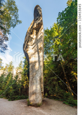 Adrspach-Teplice Rocks, sandstone formations in Hradec Kralove Region in Czech Republic, Europe. A towering rock formation rises majestically amidst a verdant forest, bathed in warm, dappled sunlight. 130453645