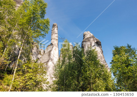 Adrspach-Teplice Rocks, sandstone formations in Hradec Kralove Region in the Czech Republic, Europe. Striking rock formations rise against a clear blue sky, framed by lush green trees on a sunny day. 130453650