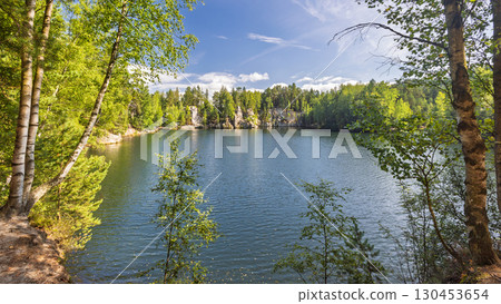 The Piskovna lake in Adrspach-Teplice Rocks area, in Hradec Kralove Region in the Czech Republic, Europe. Scenic lake surrounded by lush green forest and sandstone formations under a bright blue sky. 130453654